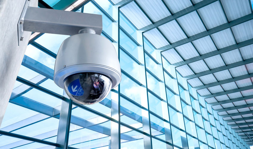 Telecamera dome bianca su staffa a parete in una galleria con vetrate e travi in acciaio sotto il cielo azzurro